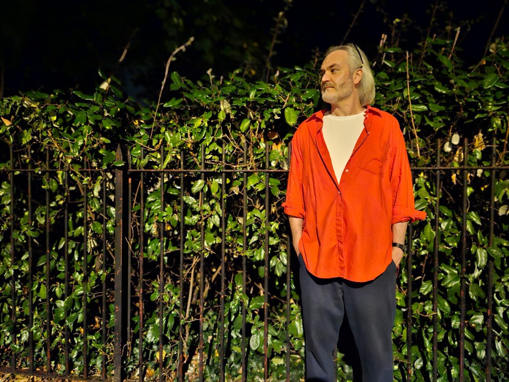Samsung Galaxy S23 Ultra sample image Outdoor Night Portrait of a man with shoulder length white hair and short beard wearing a white t-shirt and red shirt over it, standing with his hands in his pockets